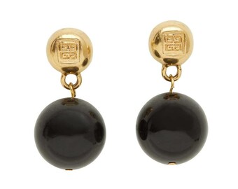 May include: A pair of gold-coloured earrings. Each earring has a square logo at the top and a large, black, spherical bead hanging below. The earrings are set against a white background.