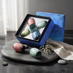 Peut inclure: Un ensemble de six tasses à thé en céramique dans une boîte cadeau bleue. Les tasses sont de différentes couleurs, dont rose, vert et bleu, avec un motif tacheté. Deux tasses sont hors de la boîte sur une pierre grise.