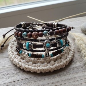 Peut inclure: Plusieurs bracelets en cuir avec des détails argentés, des perles turquoise et des perles en bois. L'un des bracelets est orné d'un pendentif tête de taureau argenté avec une pierre turquoise. Ils reposent sur un dessous de verre beige crocheté.