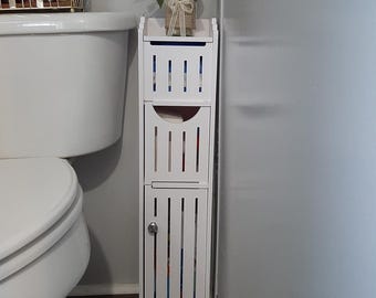 May include: A white, narrow bathroom storage cabinet with three sections. The top section has a shelf with a glass vase of flowers. The cabinet is next to a white toilet. The cabinet has vertical slats and a small door.