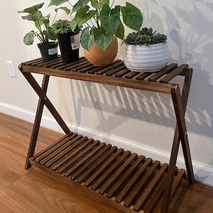 May include: A dark brown wooden plant stand with two slatted shelves. The stand holds several potted plants with green foliage, including a large-leafed plant in a terracotta pot and a succulent in a white ceramic pot.