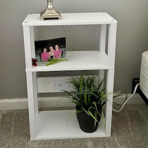 May include: A white bedside table with three shelves, a lamp, and a plant. The lamp has a silver base and a patterned shade. A framed photo and a small plant are on the middle shelf. The table includes a power outlet. The table is approximately 60 cm tall.