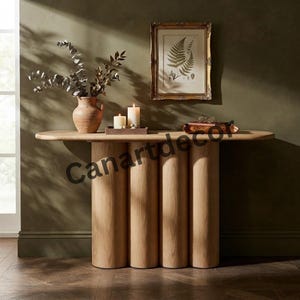 May include: A light oak console table featuring a smooth, elongated oval top supported by four thick, cylindrical vertical columns. The table is positioned against a dark olive wall, displaying a terracotta vase with eucalyptus branches.