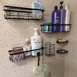 May include: Black metal shower shelves and soap holders fixed to a speckled wall. The shelves contain bottles of shampoo, body wash, and skincare products. A razor and loofah are also visible.