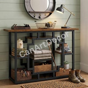 Peut inclure: Une console rectangulaire avec un plateau en bois foncé et une structure géométrique en métal noir. L'unité comporte plusieurs étagères ouvertes contenant un pot en céramique blanche, des livres empilés, des textiles gris pliés et un panier.