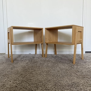 Peut inclure: Deux tables de chevet en bois clair. Chaque table a un plateau rectangulaire avec une étagère ouverte en dessous. Les tables ont quatre pieds fins et un petit détail rectangulaire sur le côté. Les tables sont posées sur un tapis marron.