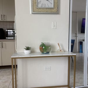 May include: A white and gold console table with a marble top, holding a small fishbowl, a potted plant, and a decorative item. A square clock with Roman numerals hangs on the wall above the table.