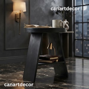 May include: A black wooden side table with a circular top and a lower shelf. The top holds an open book and a light-colored ceramic mug. The lower shelf contains a small gold-toned lamp and two stacked books. The text canartdecor is visible.