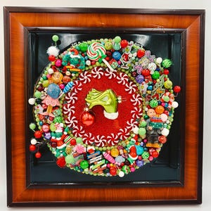 Pode incluir: Uma moldura de madeira quadrada contendo uma guirlanda de Natal circular feita de contas coloridas, figuras em miniatura, ornamentos de doces e espirais de menta. Uma mão verde central segura uma bola vermelha contra um fundo de glitter vermelho.