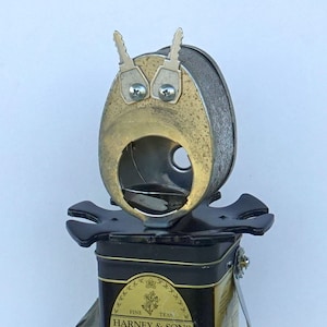May include: A whimsical sculpture made from repurposed metal objects. The head is formed from a metal container with key-shaped antennae and key-shaped eyes. The body is a black tea tin labeled "HARNEY & SONS JASMINE Floral Tea".