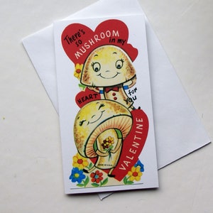 K&ouml;nnte beinhalten: Eine Valentinstagskarte mit zwei l&auml;chelnden Cartoon-Pilzen. Die Karte hat ein rotes Herz mit dem Text "There's so mushroom in my heart for you Valentine." Die Karte liegt auf einem wei&szlig;en Umschlag.