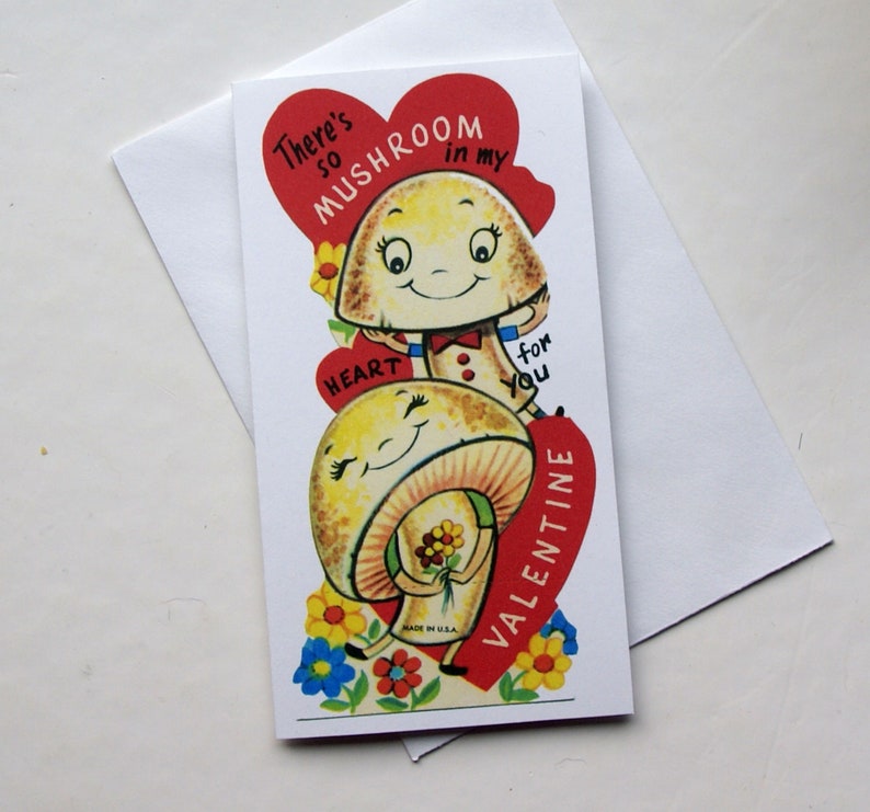 K&ouml;nnte beinhalten: Eine Valentinstagskarte mit zwei l&auml;chelnden Cartoon-Pilzen. Die Karte hat ein rotes Herz mit dem Text "There's so mushroom in my heart for you Valentine." Die Karte liegt auf einem wei&szlig;en Umschlag.