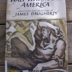 May include: A hardcover book titled Walt Whitman's America, Selections and Drawings by James Daugherty. The cover features a charcoal-style illustration of a reclining figure with a beard, resting in a landscape with mountains and an axe.