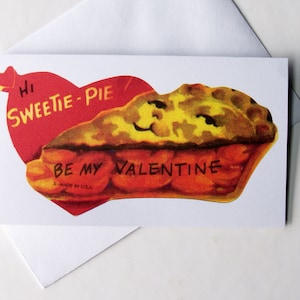 May include: A Valentine's Day card featuring a slice of pie with a smiling face, set against a red heart. The card reads "Hi Sweetie-Pie" and "Be My Valentine". The card is on a white background with a white envelope.