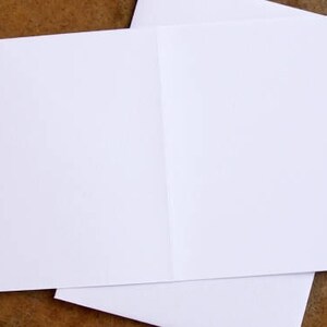 K&ouml;nnte beinhalten: Eine wei&szlig;e, gefaltete Gru&szlig;karte mit leerem Innenraum, die auf einer braunen Oberfl&auml;che liegt. Die Karte ist ge&ouml;ffnet und zeigt einen sauberen, leeren Raum f&uuml;r eine pers&ouml;nliche Nachricht. Das Papier wirkt glatt und knitterfrei.