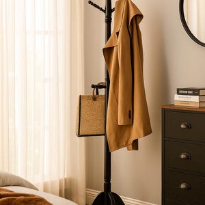May include: A black wooden coat rack with multiple hooks, holding a tan coat and a woven tote bag. The coat rack is in a bedroom setting, with a window and a dresser visible in the background.