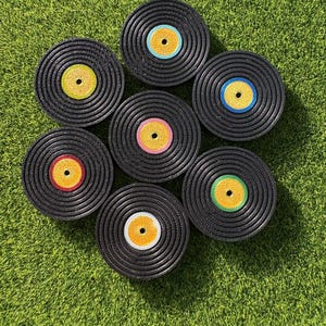May include: Seven black vinyl record coasters with colourful centre labels in yellow, red, pink, green, and blue. The coasters are arranged on a green, grassy surface.