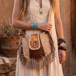 May include: A tan leather crossbody bag with fringe and a decorative flower. The bag has a brown leather panel and is adorned with silver studs. The person is wearing turquoise bracelets and necklaces.