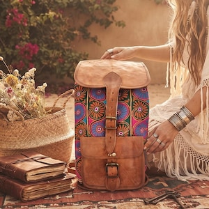 May include: A backpack featuring a tan leather top flap and front pocket with a central buckle strap. The main body is made of a vibrant fabric with a repeating pattern of blue, pink, and green circular motifs. The bag has a structured rectangular shape.