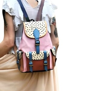 May include: A pink, brown, and blue backpack with a flap closure and adjustable straps. The flap features a white background with black polka dots. The backpack has two front pockets and brown buckles.