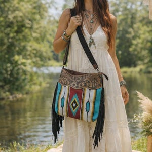 May include: A shoulder bag with a Southwestern design and black fringe. The bag has a brown leather top with floral tooling, a patterned front with red, turquoise, and white stripes, and a black strap. The bag is carried over the shoulder.