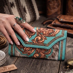 May include: A rectangular wallet featuring a turquoise leather flap with intricate floral tooling and a brown and white cowhide body. The wallet has a pointed envelope-style closure with decorative stitching along the edges and a visible brand tag.