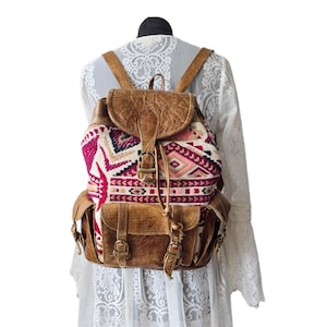 May include: A brown leather backpack with a colorful patterned fabric front. The backpack has adjustable straps, a flap closure, and side pockets. The fabric design includes pink, white, and red geometric shapes.