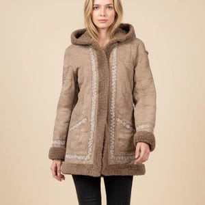 May include: A tan hooded coat with a faux fur trim and embroidered detailing. The coat features a button closure and pockets. The person in the image is wearing black pants and brown boots.