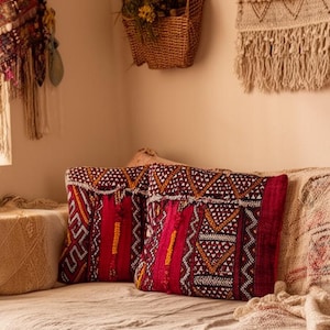 Puede incluir: Un espacio de vida acogedor con un sofá cama de tonos neutros adornado con cojines estampados en tonos rojos, naranjas y blancos. Una alfombra tejida y tapices de pared se suman a la estética bohemia. La habitación está bañada por la luz natural.