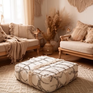 May include: A square, off-white ottoman with a geometric pattern in gray and cream. The ottoman has a textured surface and is surrounded by neutral-toned furniture and decor, creating a cozy, bohemian living space.