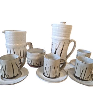 May include: A handmade ceramic coffee set in a neutral color palette. The set includes a tall coffee pot with a lid, a creamer, two small bowls, and three coffee cups with saucers. Each piece has a textured finish and dark brown linear accents.