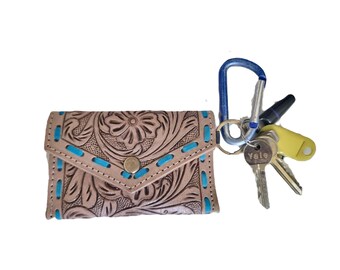 May include: A tan leather key case with an embossed floral pattern and bright blue stitching along the edges. The case features a triangular flap with a metal snap closure and is attached to a blue carabiner holding several metal keys and a yellow tag.