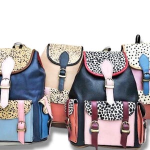 May include: A selection of colourful backpacks with a distinctive spotted flap design. Each bag features a different colour scheme, including blue, pink, and black, with contrasting straps and buckles. The bags are made of a leather-like material.