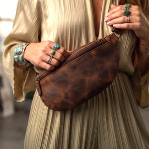 May include: A brown leather fanny pack with a smooth, distressed texture and a dark brown zipper closure. The bag features a curved, crescent-like shape and is constructed from a single piece of leather with visible natural grain and markings.