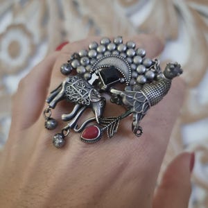 Peut inclure: Bague argentée ornée d'un éléphant et d'un oiseau, ornée d'une pierre noire carrée et d'une pierre rouge en forme de goutte. La bague présente des détails complexes et des embellissements pendants, pour un style artistique unique.