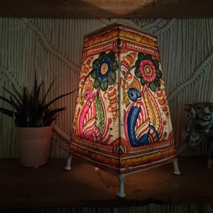 May include: A decorative lamp with a colourful, hand-painted design featuring birds and floral patterns. The lamp has a square base and a tapered shape, with white legs. The lamp is lit, casting a warm glow.