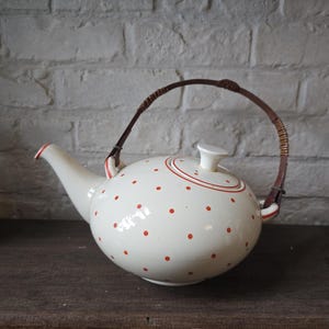 Puede incluir: Tetera de cerámica color crema con lunares rojos y un asa trenzada marrón. La tetera tiene un pico curvo y una tapa con un pequeño pomo blanco. Una banda roja rodea la tapa y la base de la tetera.