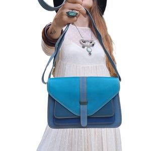 Könnte beinhalten: Blaue Leder-Umhängetasche mit Klappenverschluss. Die Tasche hat eine Kombination aus hellen und dunklen Blautönen mit einem vertikalen Riemendetail. Sie hat einen Schulterriemen und eine rechteckige Form.