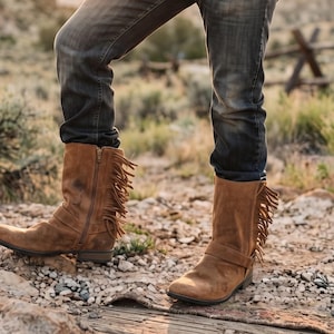 May include: Mid-calf brown suede boots featuring a side zip closure, a horizontal strap across the ankle, and a decorative fringe detail along the outer side of the shaft. The boots are worn with dark grey denim jeans.