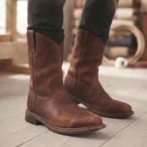 Op de afbeelding: Een paar halflange cowboylaarzen van bruin leer met een ronde neus, een blokhak en subtiele decoratieve stiksels op de schacht. De laarzen hebben een verweerde afwerking en worden gedragen met een donkergrijze spijkerbroek.