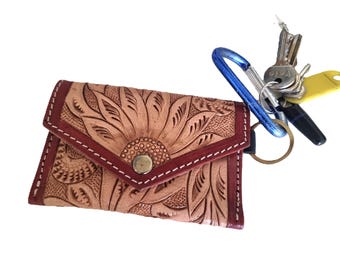 May include: A tan leather key wallet with a sunflower design, burgundy trim, and a snap closure. A blue carabiner clip holds a set of keys with a yellow tag. The wallet is approximately 4 inches wide.