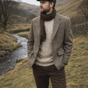 Puede incluir: Un blazer de tweed marrón, una bufanda a juego y un suéter de punto trenzado. El conjunto se completa con pantalones marrones y una gorra. El hombre está al aire libre cerca de un arroyo. El blazer tiene dos bolsillos.