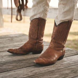 May include: A pair of mid-calf brown leather cowboy boots featuring intricate tonal stitching patterns on the shaft and a rounded toe design. The boots are worn with light-coloured trousers, showcasing a classic pull-on style with a stacked heel.
