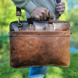 May include: A brown leather briefcase with a top handle and a shoulder strap. The bag has a textured surface and a front pocket. The briefcase is held by a person in front of a blurred green background.