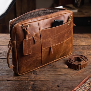 Könnte beinhalten: Eine hellbraune Leder-Laptoptasche mit Reißverschluss, einem seitlichen Tragegriff und einem abnehmbaren Schultergurt. Die Außenseite verfügt über eine Vordertasche, in der sich ein schwarzes Smartphone und ein goldener Stift befinden.