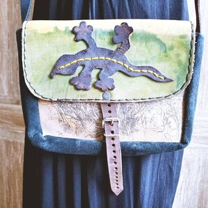 May include: A handmade leather crossbody bag with a lizard applique. The bag features a green and cream-colored flap with a dark blue lizard design stitched with yellow thread. The bag has a brown leather strap and buckle.