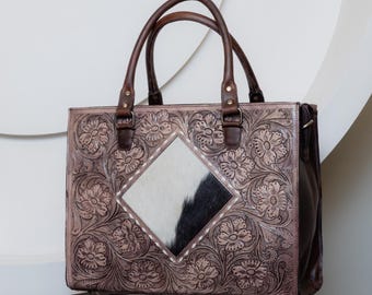Puede incluir: Bolso tote de cuero marrón con detalles florales en relieve. El bolso presenta un panel en forma de diamante de piel de vaca en blanco y negro. Tiene dos asas de cuero marrón y un diseño estructurado.