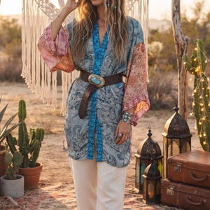 May include: A long-sleeved kimono-style tunic featuring a blue and grey paisley pattern with contrasting peach-coloured sleeves, a bright blue trim, and a brown leather belt with a large oval turquoise-coloured buckle worn over light-coloured trousers.