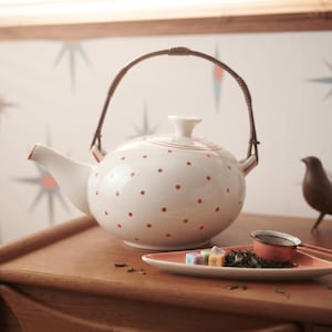 Può includere: Una teiera in ceramica bianca a pois rossi con un manico marrone ricurvo, accompagnata da un piattino triangolare contenente foglie di tè sfuse, piccoli cubetti di zucchero colorati e un colino in metallo con bordo e manico rosso.