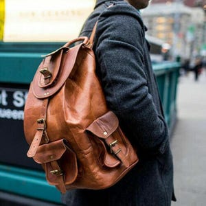 May include: A tan leather backpack featuring a large main compartment with a fold-over flap, secured by a buckle strap. The bag includes two smaller exterior pockets on the front, each equipped with a flap and a buckle closure, providing storage.